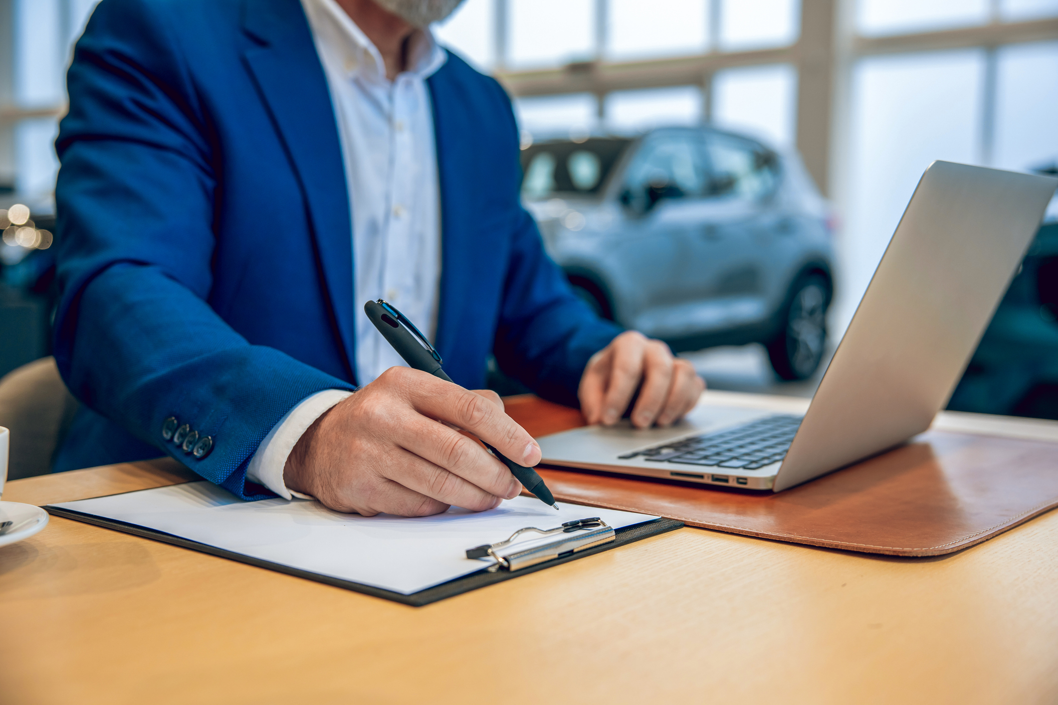 Unrecognizable man in car showroom wearing suit using laptop preparing sales agreement